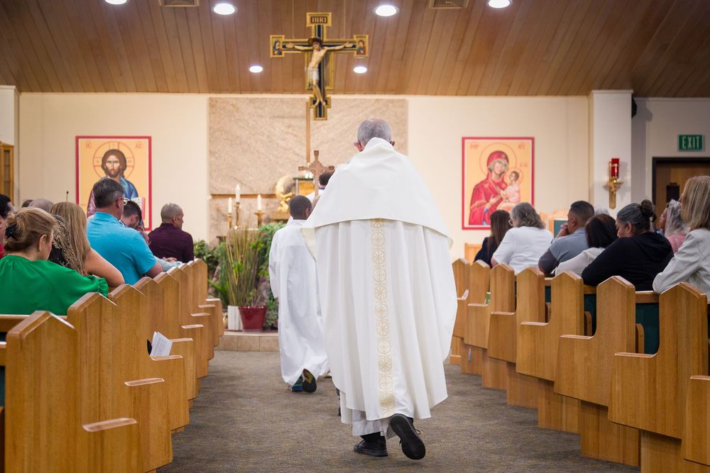 A priest in white robes walks down the church aisle during mass while parishioners sit in wooden pews.