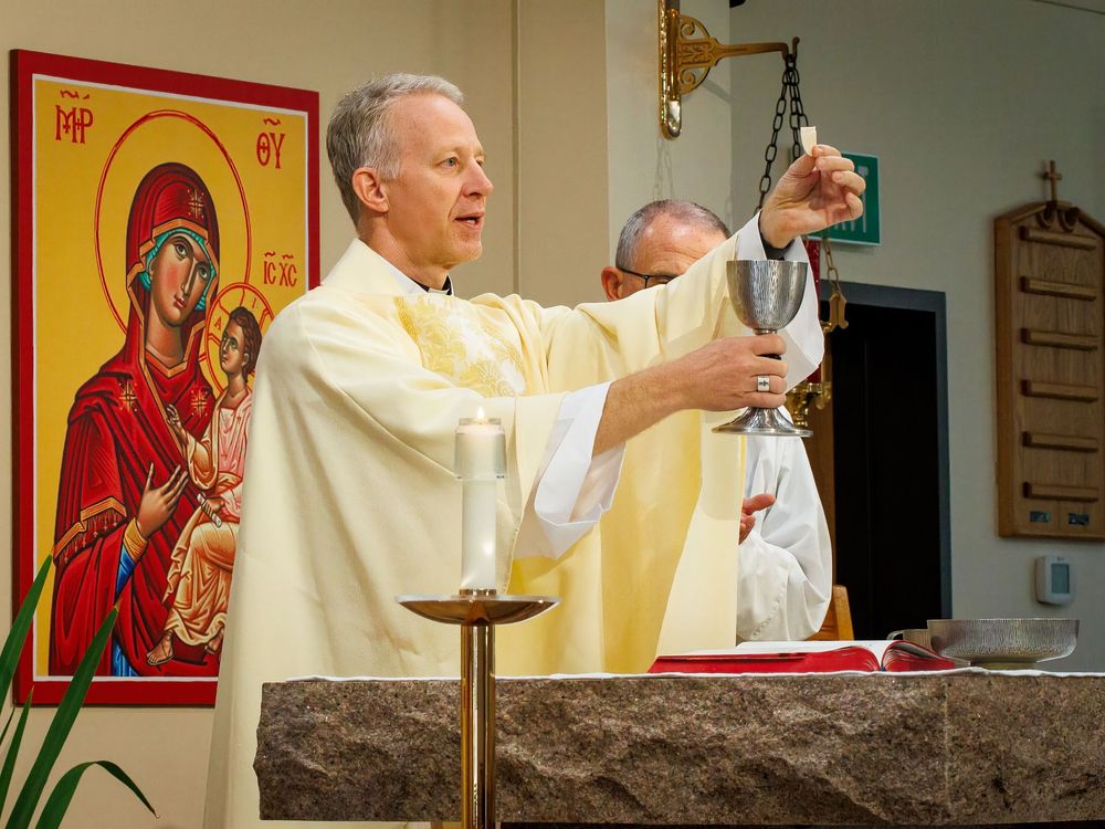 A priest in cream-colored vestments raises a ceremonial chalice at the altar during Mass with an icon visible in background.