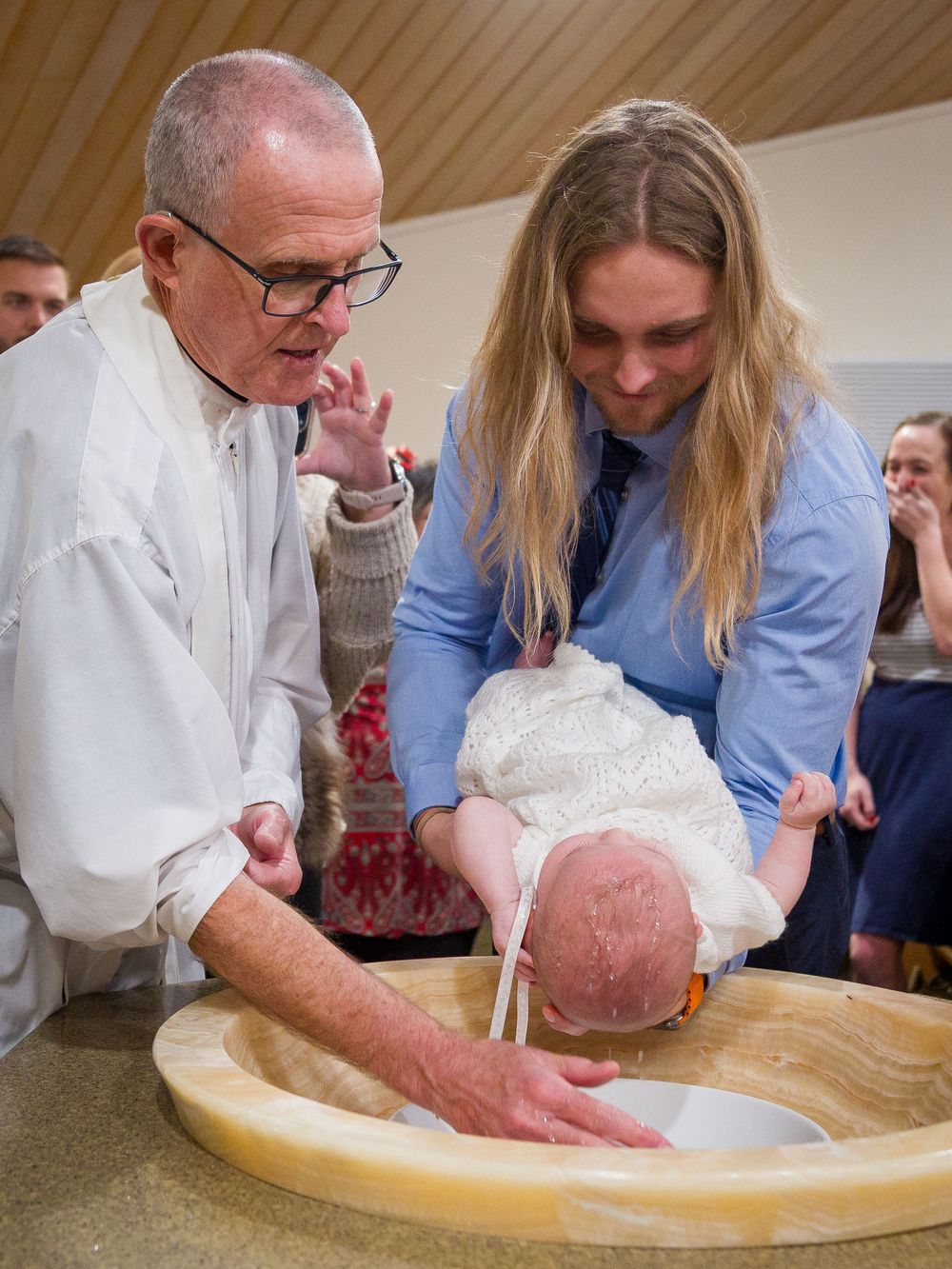Two people gather around a wooden baptismal font during a religious ceremony indoors.
