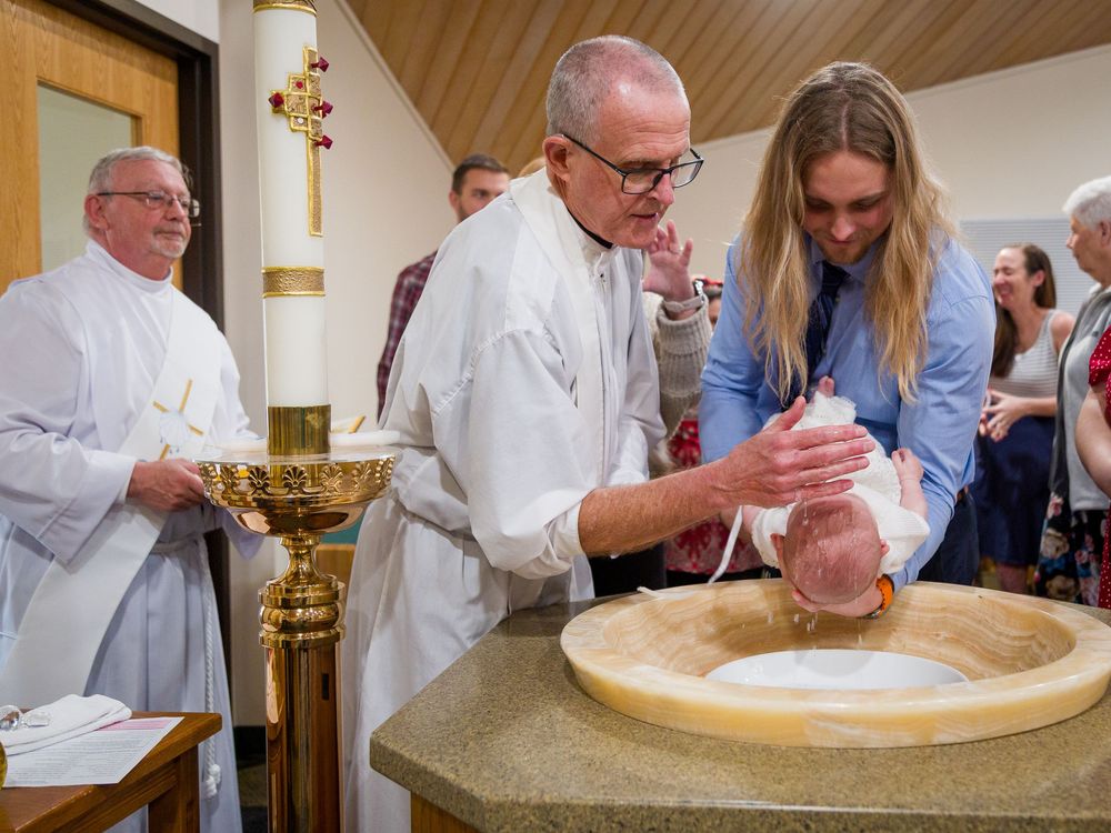 A clergy member performs a baptism at a stone baptismal font while others observe in a church setting.