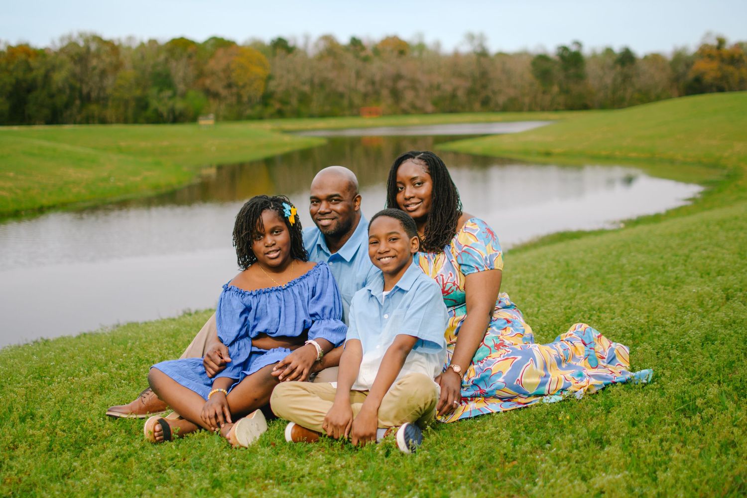 A happy family sits together on green grass near a scenic pond on a sunny day.