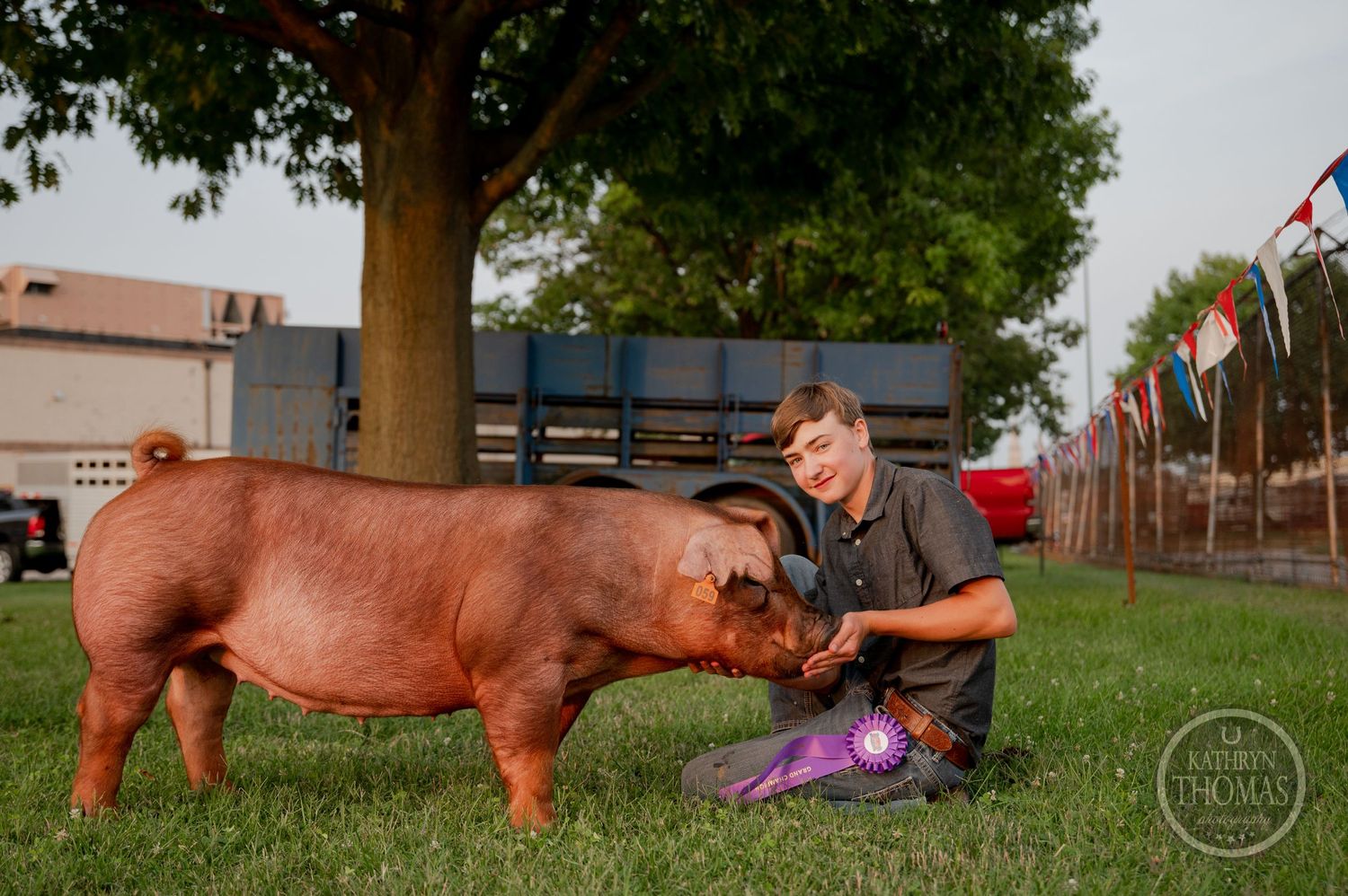 A show pig and its handler pose for a photo on grass beneath a large tree at a livestock event.