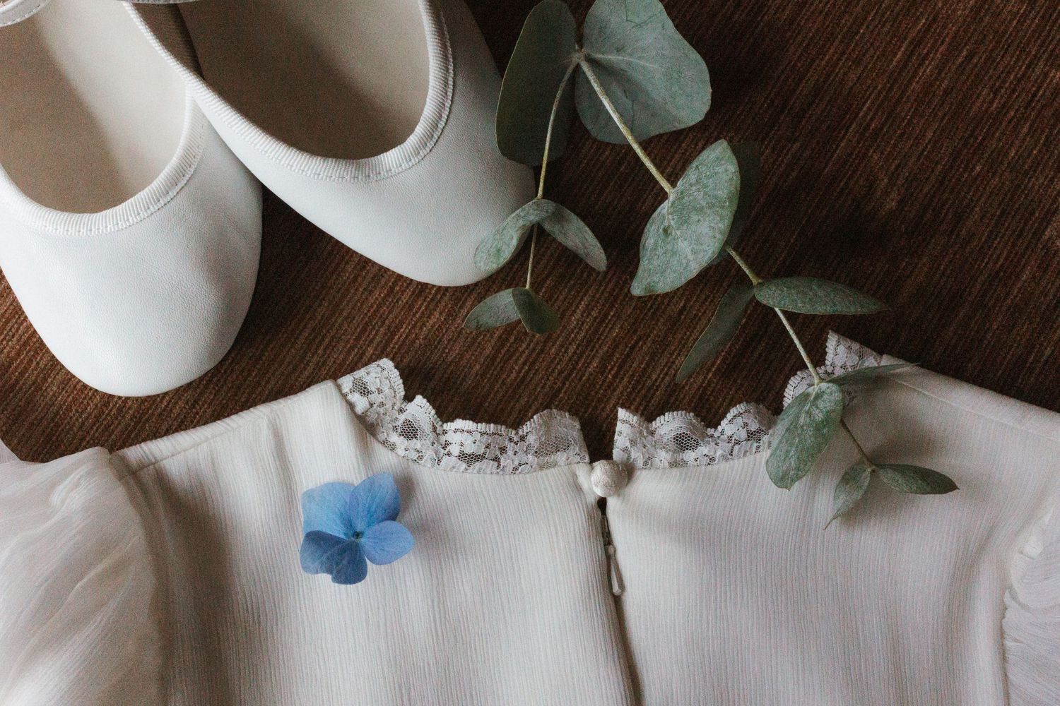 White wedding flats and dress details with blue butterflies and eucalyptus leaves on wooden surface.