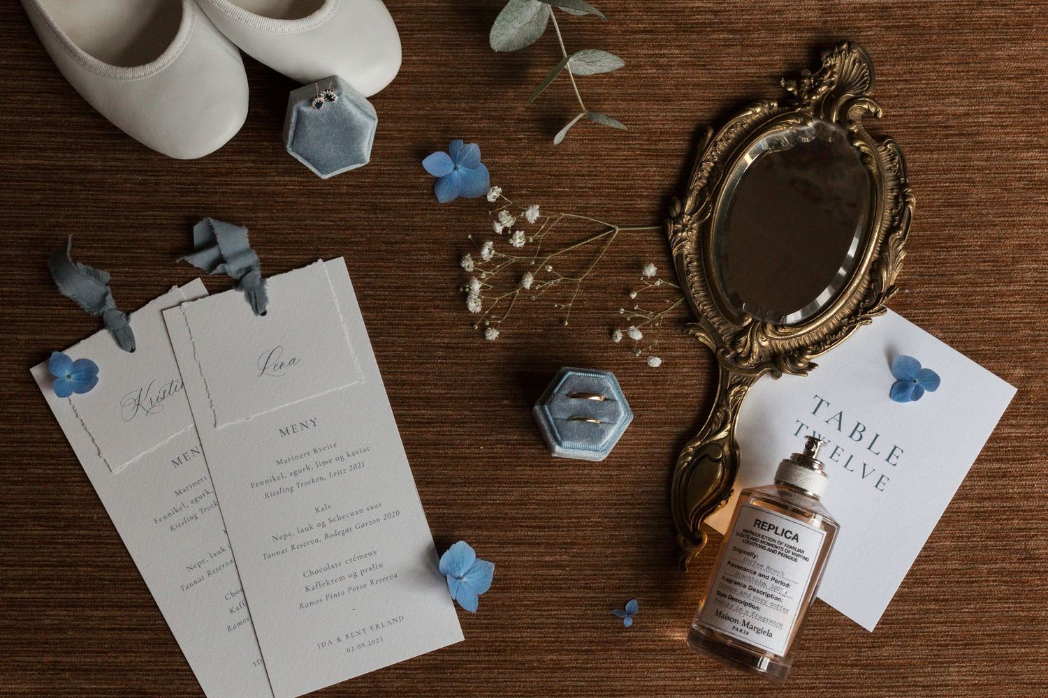 Wedding details flatlay with white shoes, invitations, perfume bottle and vintage mirror on wooden surface.