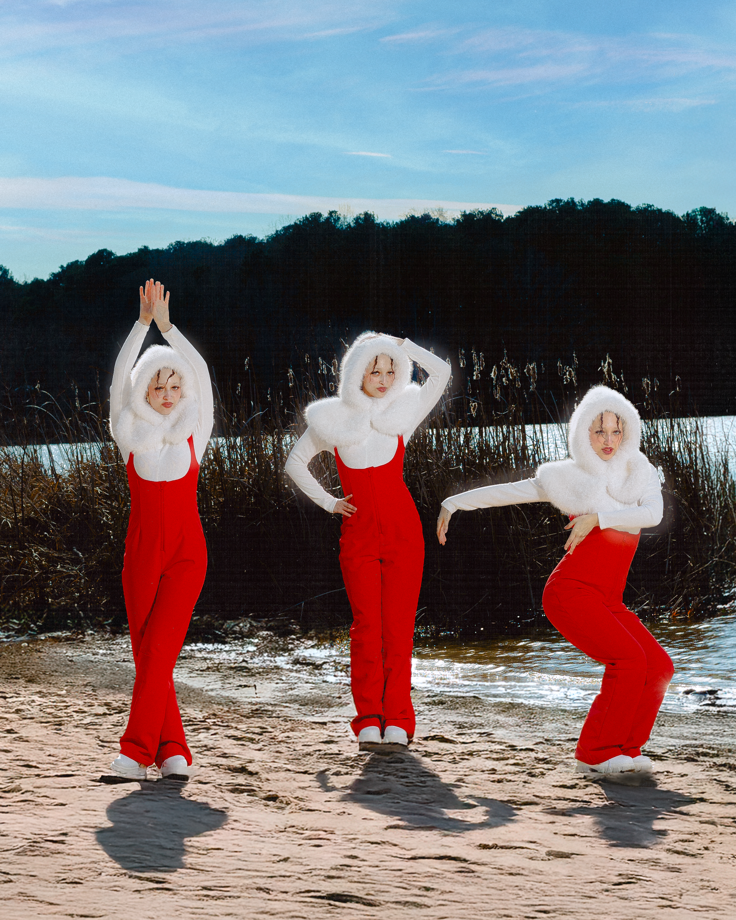 Figure in flowing red dress and white furry hood performs dynamic dance poses on a beach with trees in background.