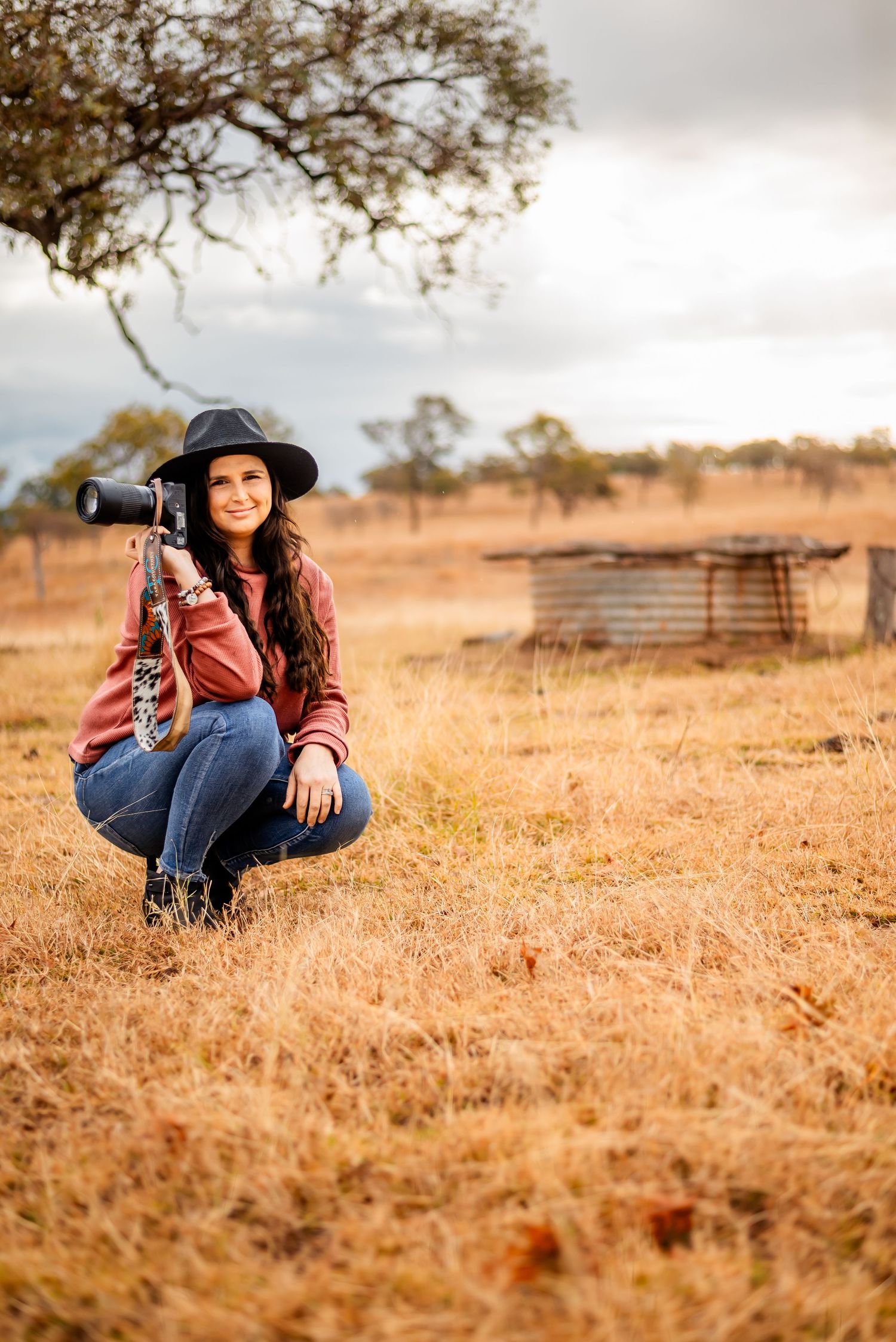 Professional photographer in hat and jeans crouches in golden field with camera during sunset.