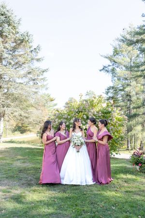 Bride in white gown surrounded by four bridesmaids in mauve dresses under a floral arch among tall trees.