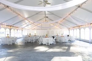 Elegant white wedding reception tent decorated with string lights, draped fabric, and white-clothed round tables.
