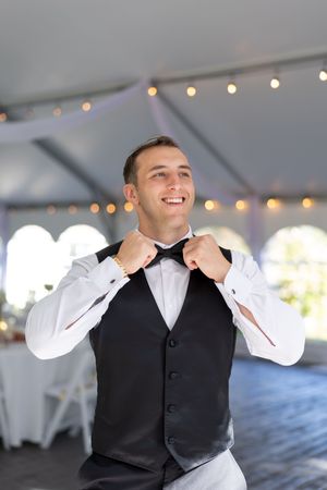 Smiling groom in black vest and bow tie adjusting his collar inside a decorated wedding reception tent.