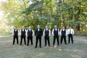 Groom with groomsmen in matching black vests and bow ties posing in a line on a grassy lawn.