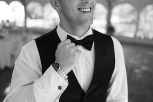 Black and white portrait of a smiling groom in a black vest adjusting his bow tie at the reception.