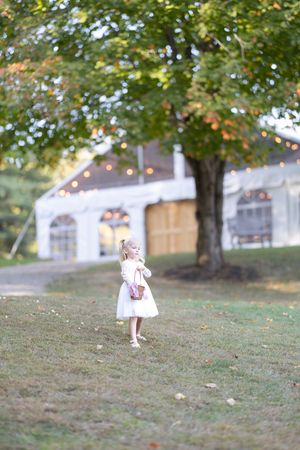 Young flower girl in white dress standing on a lawn near a large tree with a lit wedding tent behind.