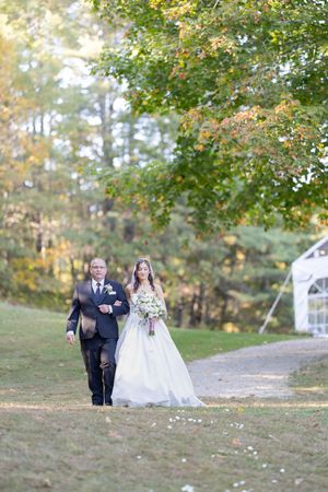 Father walking bride in a white ballgown down a petal-lined path toward a white wedding tent outdoors.