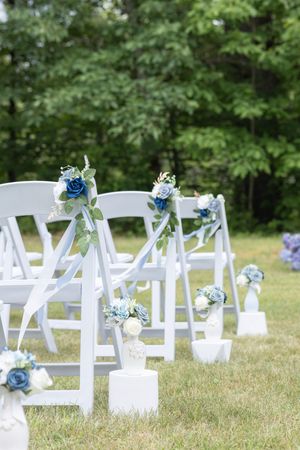 White folding chairs decorated with blue and white floral arrangements at an outdoor wedding ceremony.