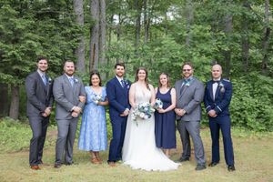 Wedding party group portrait with bride, groom, bridesmaids and groomsmen in blue and grey attire outdoors.