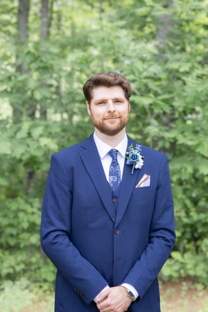 Handsome groom in a navy blue suit with floral boutonniere posing outdoors among green trees.