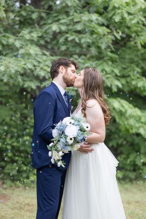 Bride and groom sharing a kiss outdoors surrounded by bubbles, bride holding a blue and white bouquet.