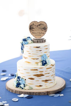 Two-tier white wedding cake with blue floral decorations and custom wooden heart topper reading Adam and Sydney.