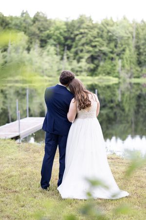 Bride and groom embracing from behind near a serene lake with a wooden dock and lush green trees.