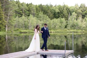 Bride and groom walking hand in hand on a wooden dock with a peaceful forest lake backdrop.