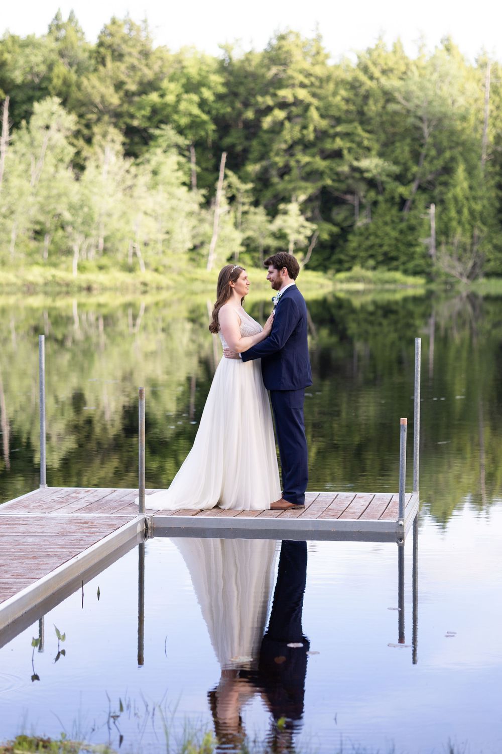 Bride and groom standing on a wooden dock sharing a romantic moment with lake reflections behind them.