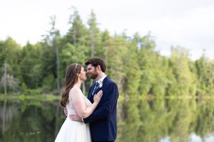 Bride and groom sharing an intimate moment by a calm reflective lake surrounded by green forest trees.