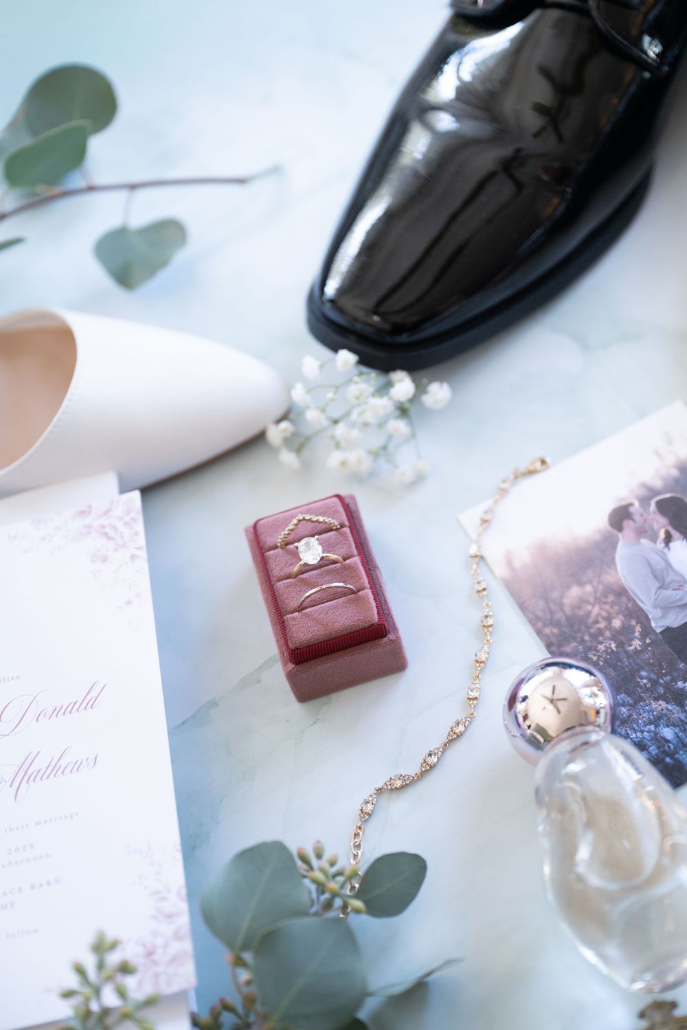 Flat lay of wedding rings in a velvet box, invitation, shoes, bracelet, and eucalyptus on marble surface.