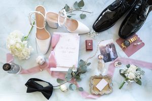 Elegant wedding flat lay with white heels, black dress shoes, bow tie, floral boutonnieres, and pink invitation suite.