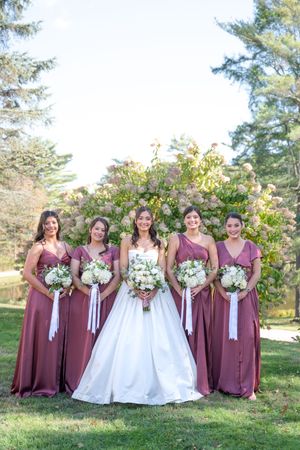 Bride in white ball gown poses with four bridesmaids in dusty rose dresses holding white bouquets outdoors.