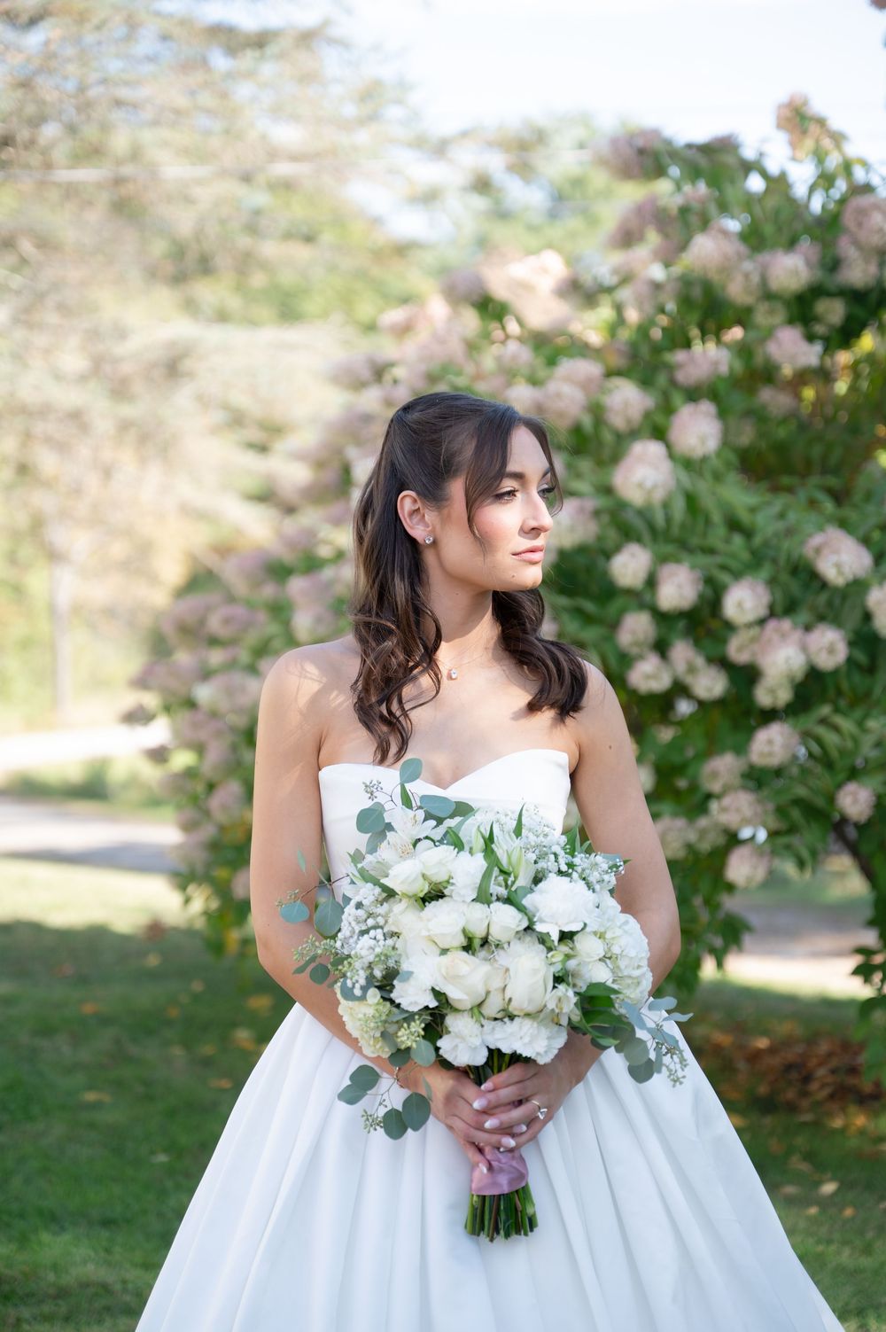 Beautiful bride in strapless white ball gown holds white and greenery bouquet before blooming pink flower trees.