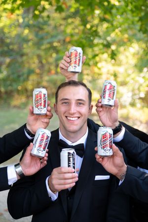Smiling groom in black tuxedo surrounded by groomsmen all holding up cans of beer outdoors.
