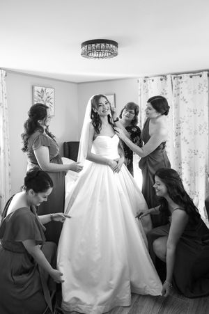 Black and white photo of bridesmaids helping bride into her white wedding gown in a bright room.