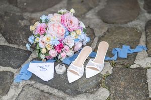 A bridal flat lay featuring white heeled mules, a colorful pink and blue bouquet, and a folded handkerchief on stone.
