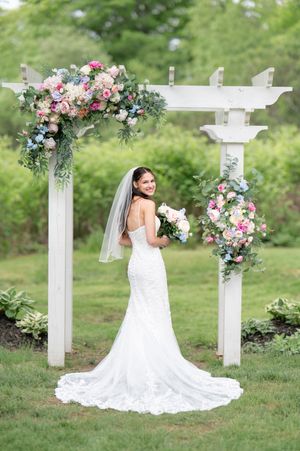 A bride in a backless lace gown and veil poses under a white floral arch with pink and blue blooms.