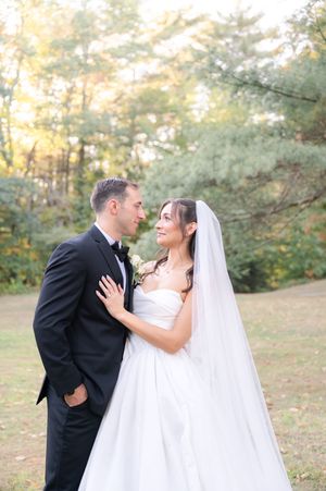 Bride in white gown with veil and groom in black tuxedo share a romantic moment in autumn woods.