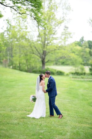 A bride and groom share a kiss on a lush green lawn surrounded by trees on their wedding day.