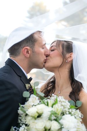 Bride and groom share a romantic kiss under the wedding veil while holding a white floral bouquet.