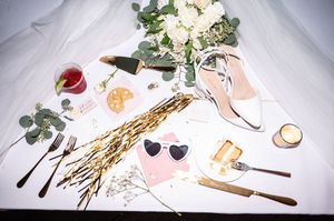 Wedding flat lay with white heels, heart sunglasses, white bouquet, gold wheat, and red cocktail drink.