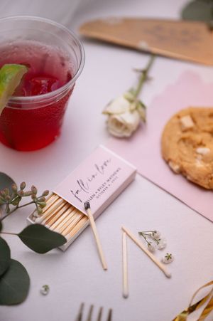 Wedding detail flat lay featuring custom matchbox, red cocktail, white rose, cookie, and eucalyptus greenery.
