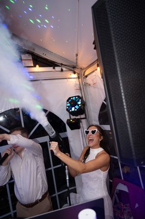 Bride in white dress and heart sunglasses shoots confetti cannon at wedding reception under tent with colorful lights.