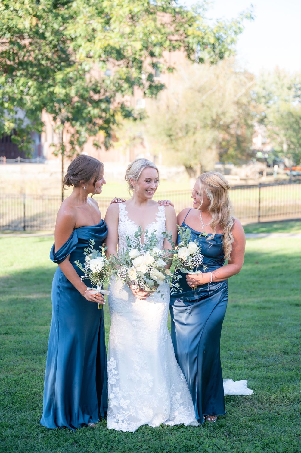 Bride in lace gown stands with two bridesmaids in blue satin dresses holding white bouquets outdoors.