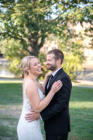 Laughing bride in lace gown embraces smiling groom in black tuxedo during outdoor wedding portrait.