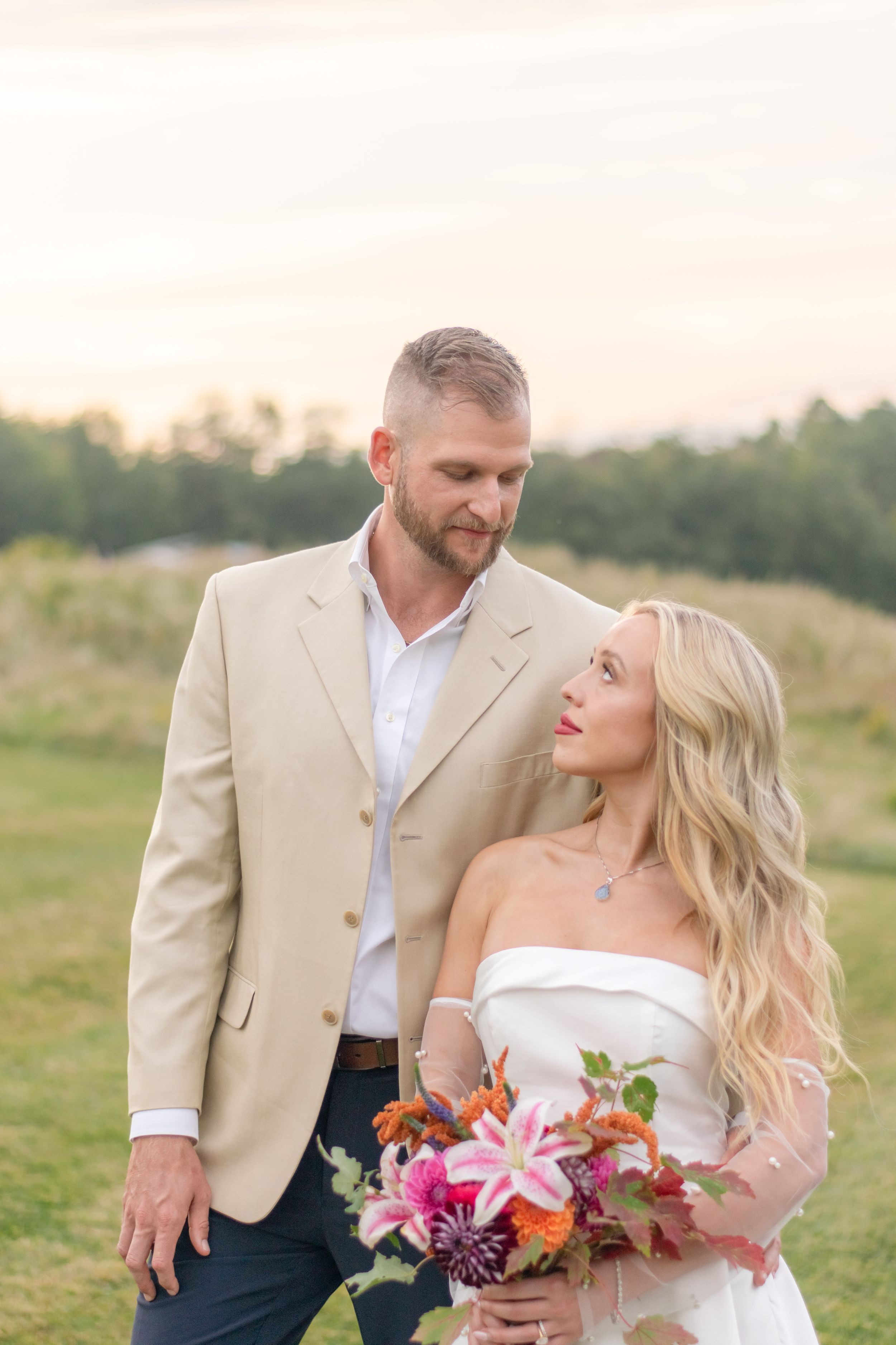 Groom in tan blazer gazing at smiling blonde bride holding colorful pink and orange floral bouquet at sunset.