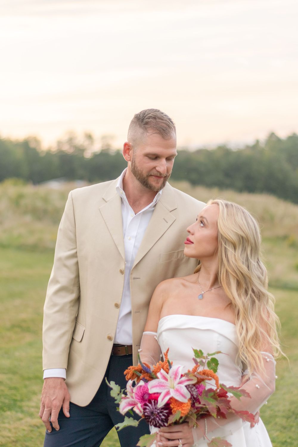 Groom in tan blazer gazing at smiling blonde bride holding colorful pink and orange floral bouquet at sunset.