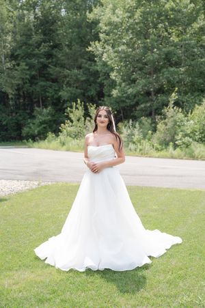 Bride in strapless white ballgown with long train stands smiling on green lawn surrounded by lush trees.