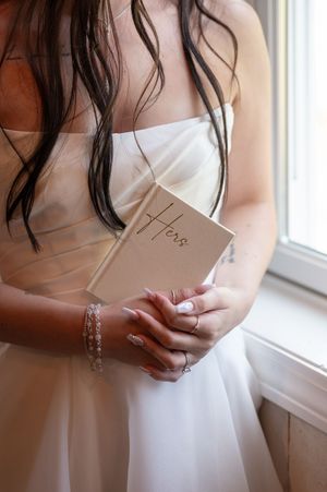 Bride in white strapless gown holds a kraft paper vow book labeled Hers near a sunlit window.