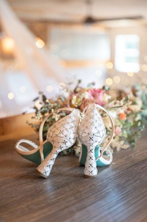 Elegant white lace wedding heels with teal soles displayed on wooden table beside a romantic floral bouquet.
