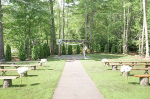 Rustic outdoor wedding ceremony setup with wooden benches lining a path leading to a decorated arbor in woods.