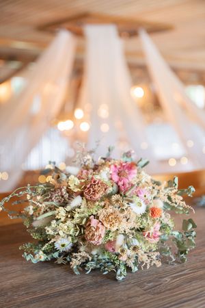 Romantic bridal bouquet of dried pink and cream wildflowers on wooden table with soft bokeh string lights behind.