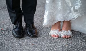 Close-up of bride's floral embellished sandals and groom's black dress shoes on pavement.